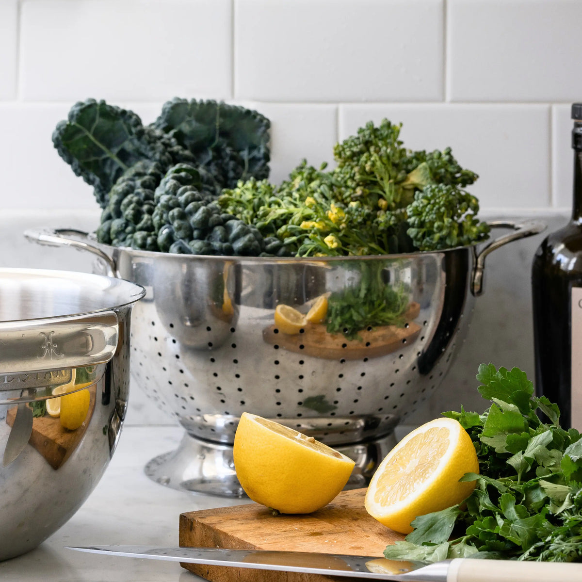 The Hestan Provisions Stainless Steel Colander, dishwasher safe and designed for superior drainage, overflows with leafy greens on the kitchen counter amid a cut lemon, chopped greens, a knife, a mixing bowl, and a bottle—perfect for fresh meal prep.