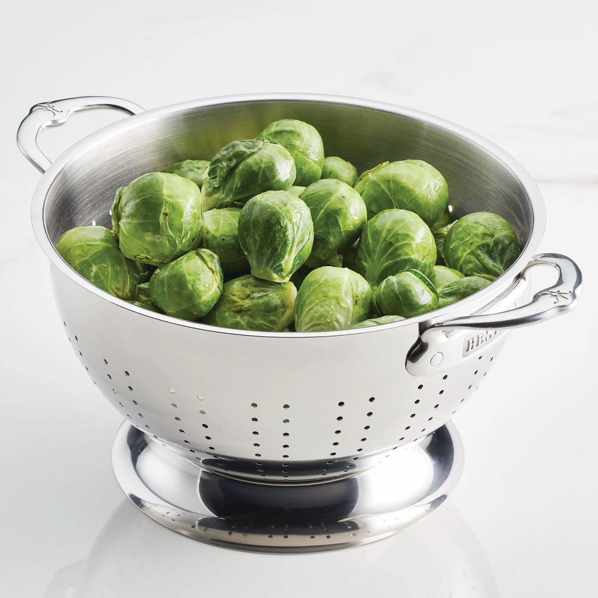 A Hestan Provisions Stainless Steel Colander with excellent drainage holds fresh Brussels sprouts and rests on a white surface.