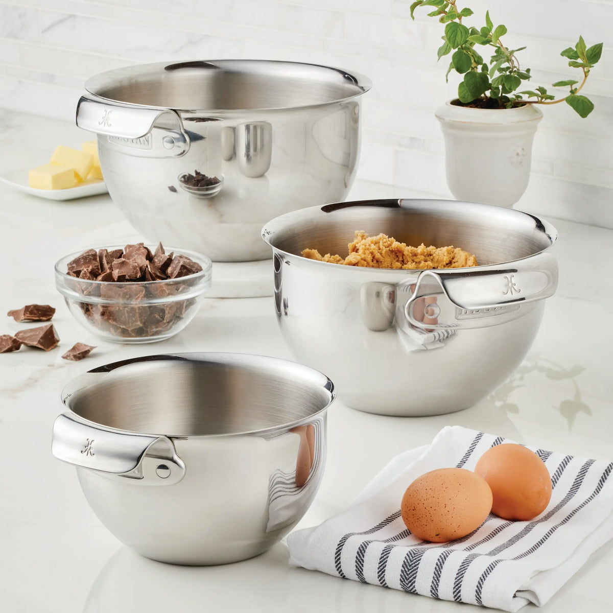Three Hestan Provisions Stainless Steel Mixing Bowl Set, 3-Piece, are on the counter—one contains brown sugar. Beside them are two eggs, a striped towel, chocolate chunks in a glass bowl, butter on a dish, and a potted plant.