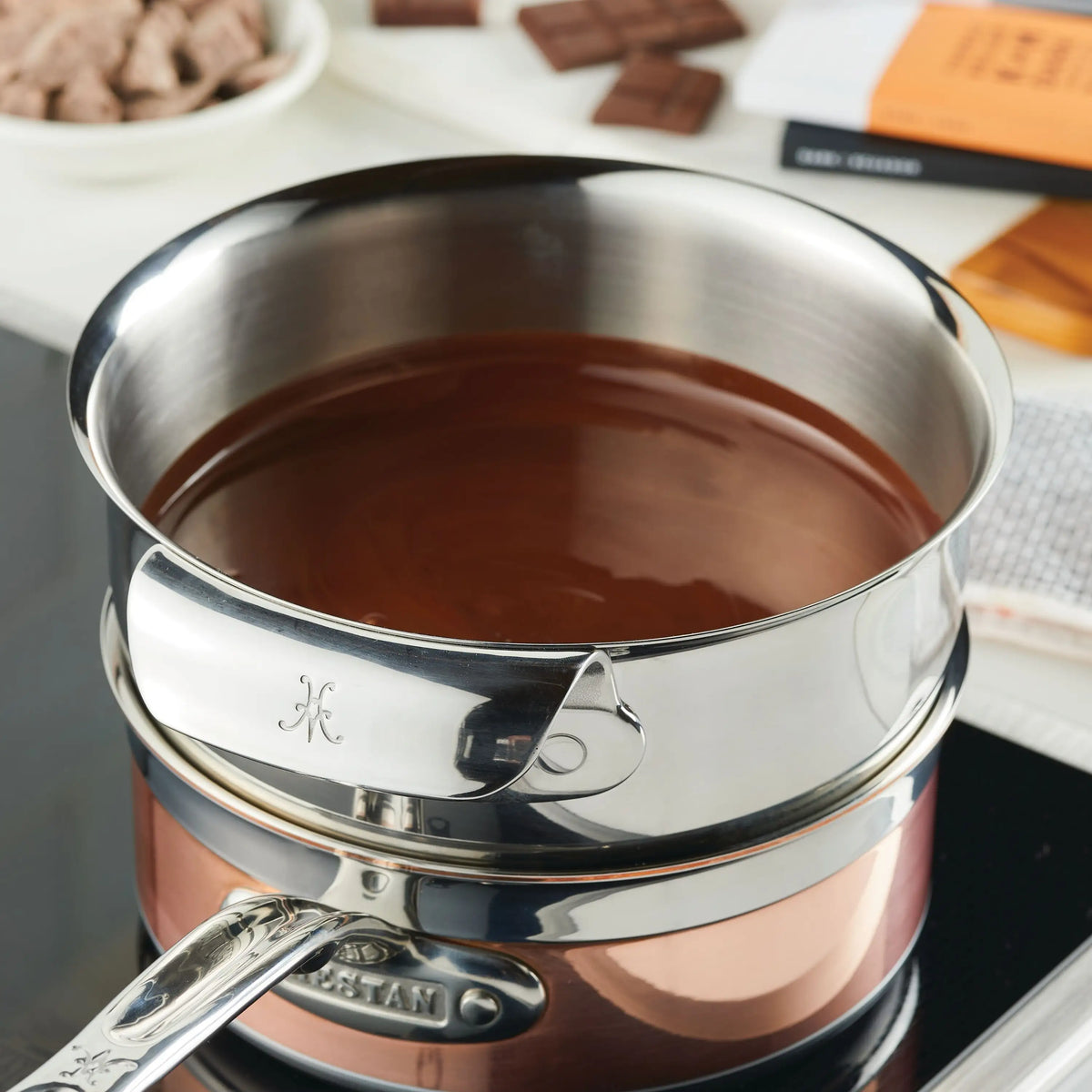 A metal saucepan melts chocolate on a stove, with Hestan Provisions Stainless Steel Mixing Bowl Set (3-Piece) nearby. Chocolate bars and a white bowl with chocolate chunks appear in the background as part of the double boiler setup.