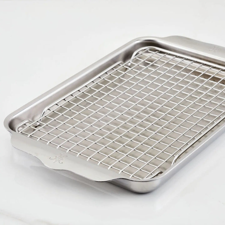 The Hestan Provisions OvenBond Tri-ply Quarter Sheet Pan with Rack, featuring small handles and a shiny stainless steel finish, sits on a white surface reflecting light beautifully.