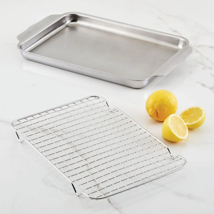 An OvenBond Tri-ply Quarter Sheet Pan with Rack by Hestan Provisions is on a white marble surface, next to a cooling rack. Two whole lemons and a lemon half are nearby.