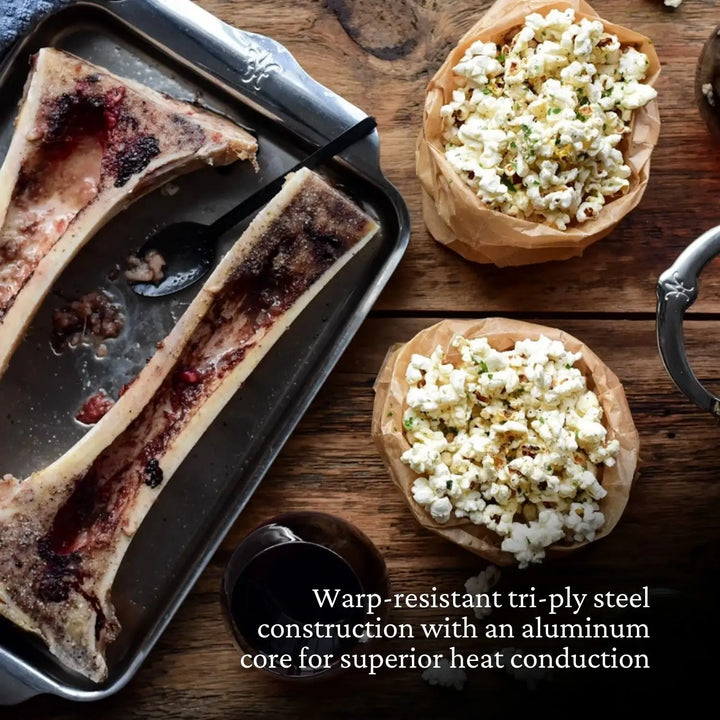 A wooden table with cooked bone marrow on a tray and popcorn in paper bowls. A dark glass is partially visible. Text reads: Hestan Provisions OvenBond Tri-ply Quarter Sheet Pan with Rack boasts warp-resistant stainless steel and an aluminum core for top-tier heat conduction, ideal for roasting.