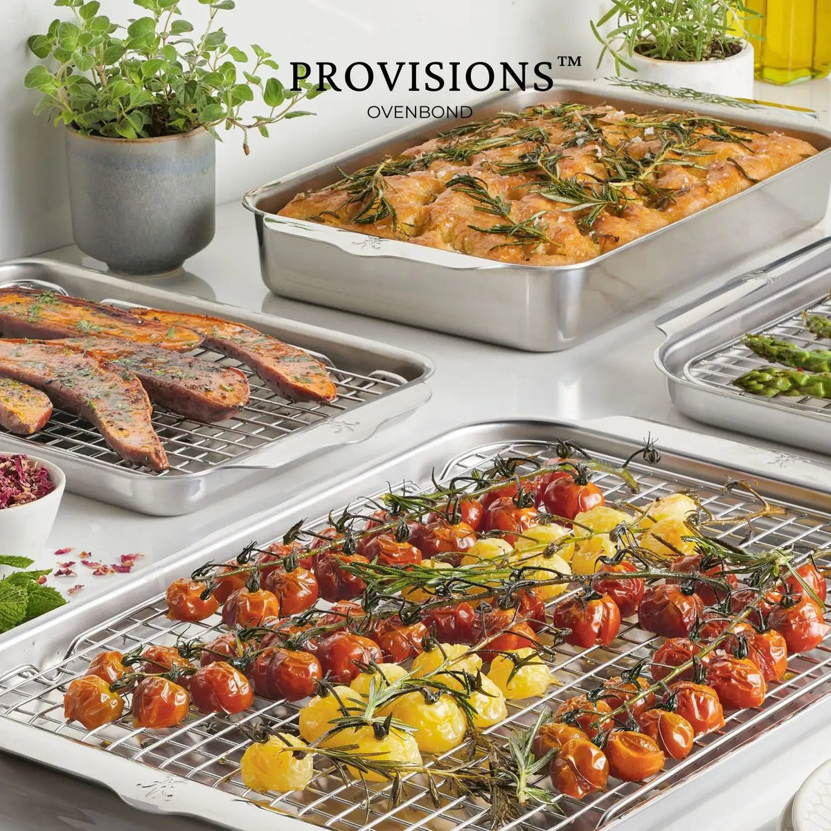 Four roasting trays, including a Hestan Provisions OvenBond Stainless Steel Quarter Sheet Pan Rack, showcase roasted tomatoes and peppers, baked rosemary bread, roasted vegetables, and cooked meat. Fresh herb pots add greenery below a backdrop highlighted by an oil bottle.