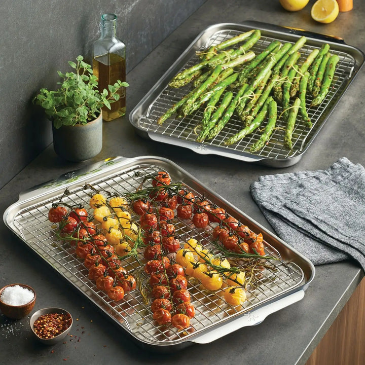 Two trays on a countertop display roasted vegetables using Hestan Provisions OvenBond Tri-ply set. Asparagus sits on one, while red and yellow vine tomatoes are on the other, all perfectly roasted on a stainless steel sheet. Nearby are a plant, oil bottle, pepper mill, salt bowl, and light gray cloth.