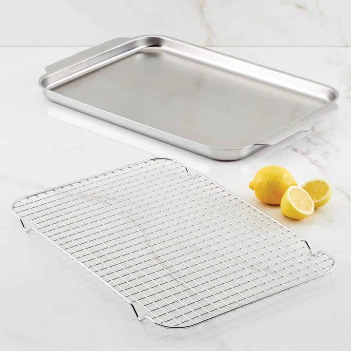 An OvenBond Tri-ply Sheet Pan and cooling rack by Hestan Provisions grace the white marble surface, complemented by two whole lemons and one sliced half, adding a splash of color to the elegant setup.