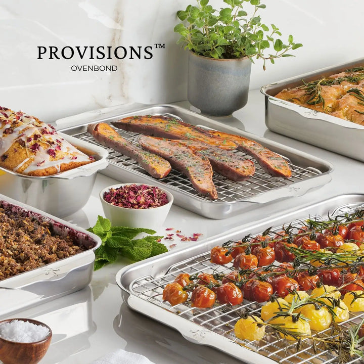 Baking trays filled with assorted foods like bread, potato wedges, and roasted tomatoes rest on the Hestan Provisions’ Stainless Steel Sheet Pan, surrounded by a pot of fresh herbs and a bowl of spices. Overhead, the OvenBond Tri-ply 4-Piece Set enhances the kitchen setup.