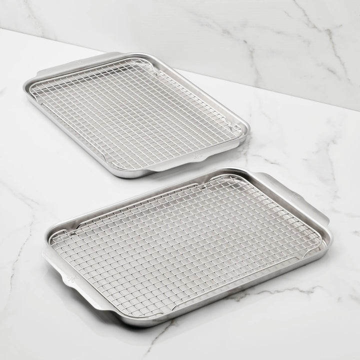 The Hestan Provisions OvenBond Tri-ply 4-Piece Set includes two stainless steel sheet pans with matching wire racks, featuring raised edges and handles for easy baking or roasting, all shown on a white marble surface.