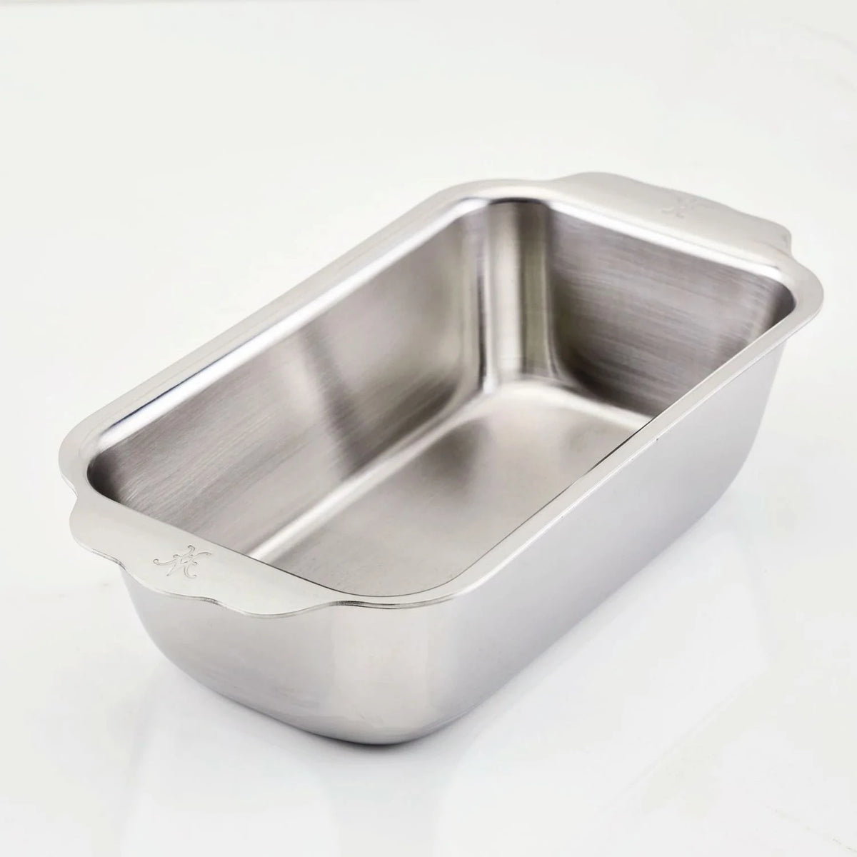 The Hestan Provisions OvenBond Tri-ply Stainless Steel 1-Pound Loaf Pan, featuring a shiny and reflective finish, rounded corners, and side handles, sits on a white surface. This warp-resistant pan combines durability with style for your kitchen.