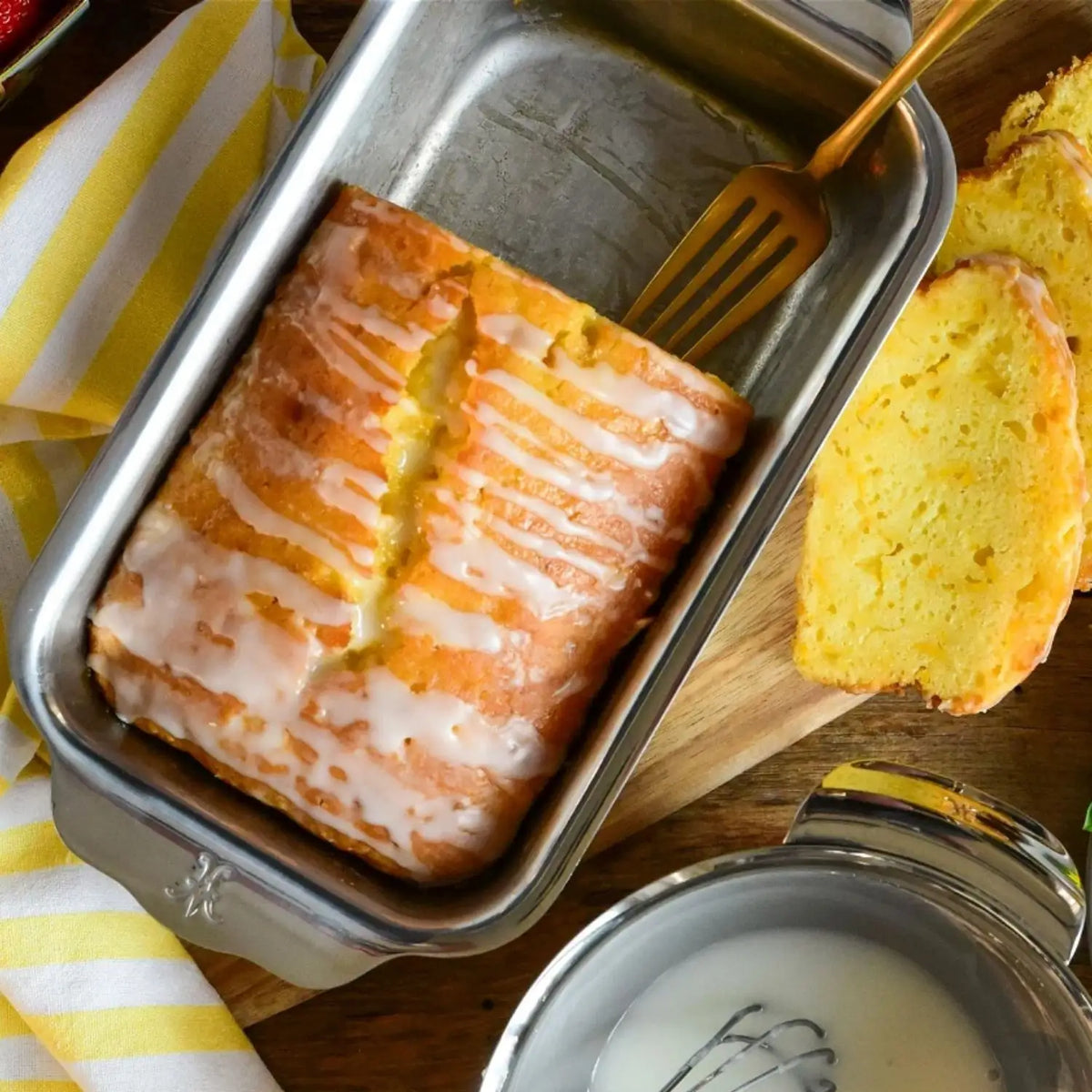 A lemon cake with icing drizzled on top sits in an OvenBond Tri-ply Stainless Steel 1-Pound Loaf Pan by Hestan Provisions. A gold fork rests inside, while sliced pieces lie on a wooden board nearby. In the foreground, theres a bowl of icing with a whisk and a yellow-striped cloth partially visible.