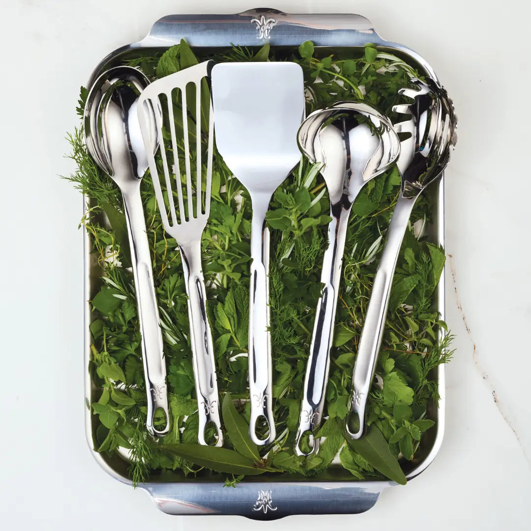 A metal tray with fresh green herbs displays Hestan Provisions Essential Chefs Tool Set, 5-piece, featuring a ladle, slotted spatula, flat spatula, serving spoon, and pasta server. These stainless steel utensils are neatly arranged.
