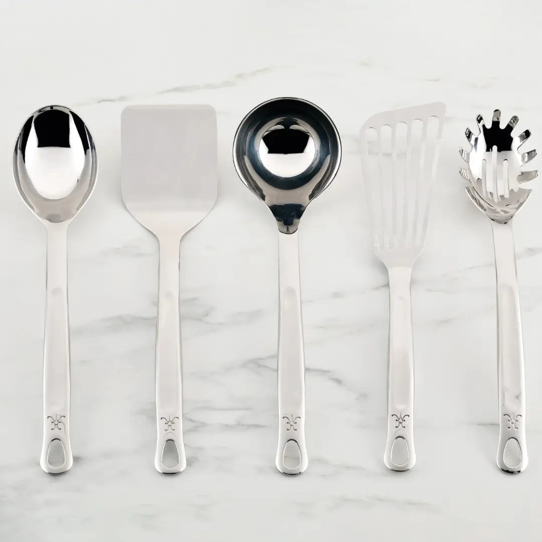 A set of five Hestan Provisions Essential Chefs Tools, including a serving spoon, flat spatula, ladle, slotted spatula, and spaghetti server, elegantly rest on a white marble surface. Each piece boasts a handle with a decorative emblem.