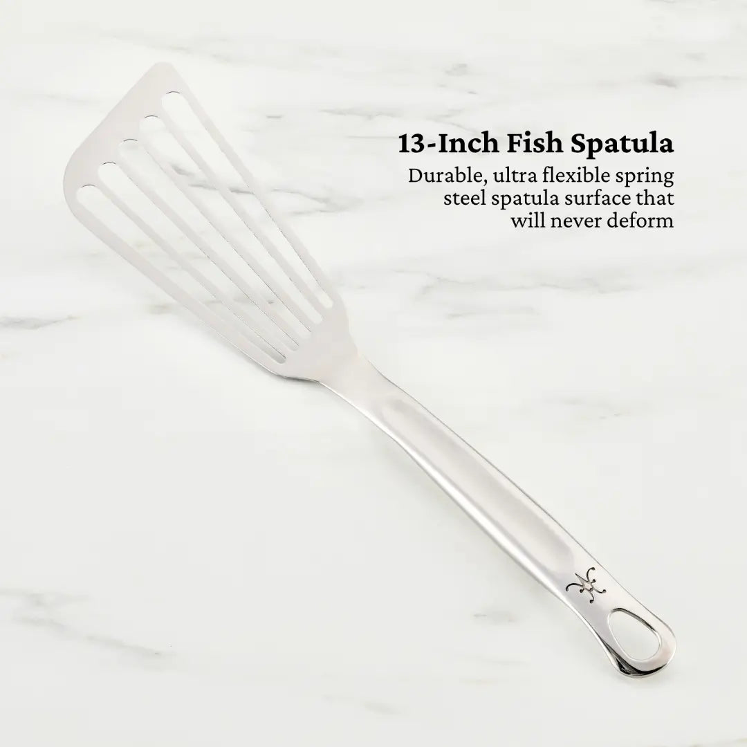 Image of a 13-inch Hestan Provisions fish spatula from the Essential Chefs Tool Set, 5-piece. It features a durable, ultra-flexible spring stainless steel surface with slotted openings and a curved handle with a decorative design, laid on marble. Text highlights its qualities.