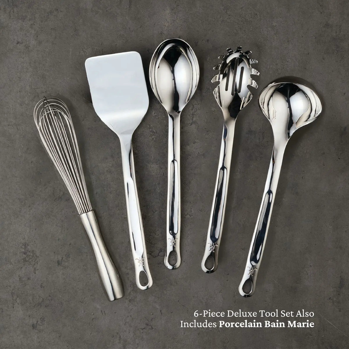A set of Hestan Provisions Deluxe Chefs Tool Set, 6-piece, consisting of stainless steel whisk, spatula, serving spoon, pasta server, and ladle is displayed on a gray surface. Text in the corner reads 6-Piece Deluxe Tool Set Also Includes Porcelain Bain Marie.