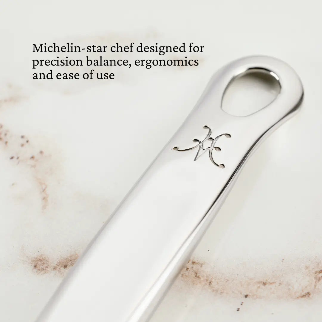 Close-up of a polished stainless steel utensil handle with an inscribed stylized letter K or H. The handle, part of the Hestan Provisions Ultimate Chefs Tool Set (8-piece), features an oval hole at the end and is designed by a Michelin-star chef for precision balance, ergonomics, and ease of use.