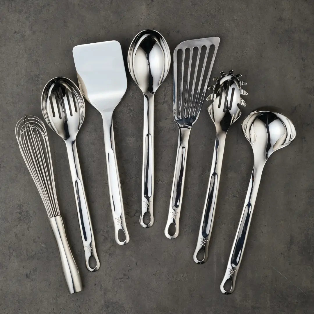 The Hestan Provisions Ultimate Chefs Tool Set, 8-piece, crafted from 18/10 stainless steel and used by Michelin-starred chefs, features tools like a whisk, slotted spoon and turner. Elegantly arranged on a gray surface, all pieces have handy hanging holes for easy storage.