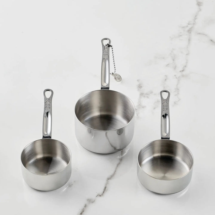 Three stainless steel measuring cups from the Hestan Provisions® Odd-Size Measuring Cups Set, 3-Piece are arranged on a white marble surface. Each handle features measurement markings, while one cup’s handle is connected to a small ball chain with a round tag—showcasing Hestan Provisions®’ dedication to functional design in their stainless steel measuring cup set.