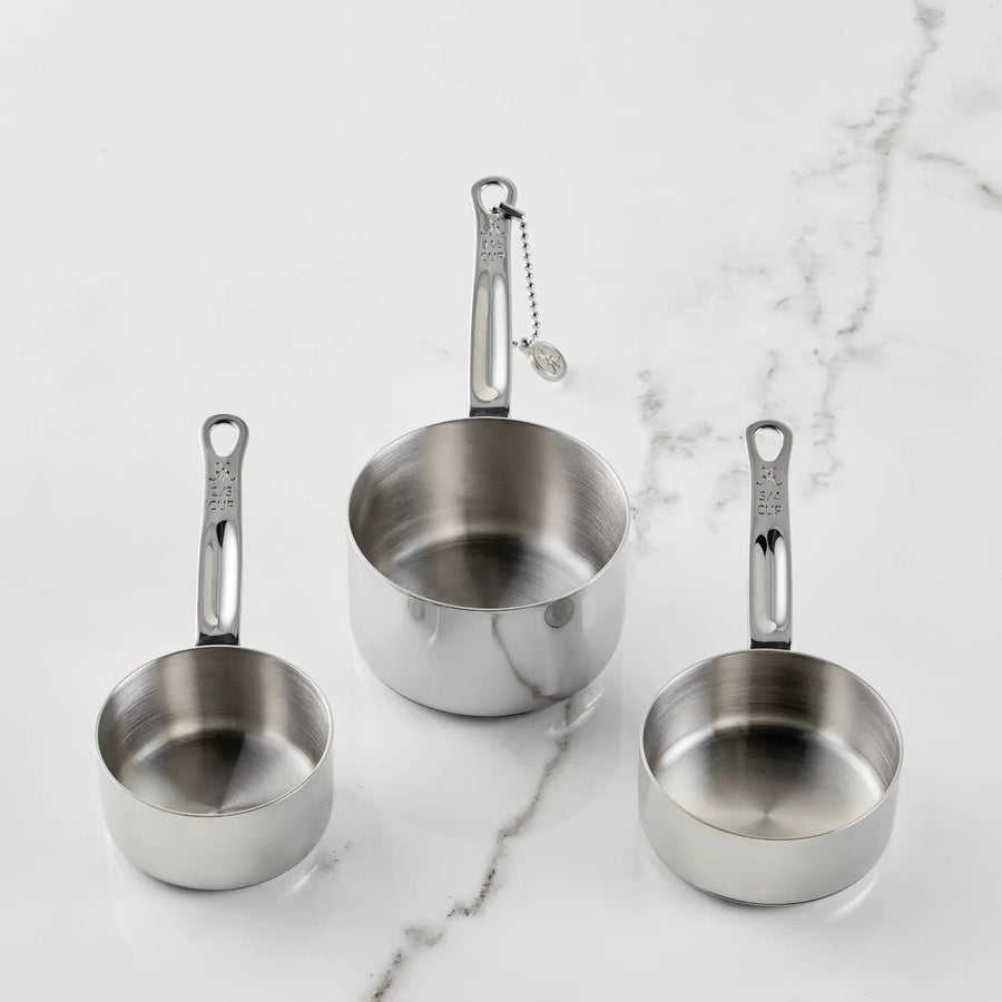 Three stainless steel measuring cups from the Hestan Provisions® Odd-Size Measuring Cups Set, 3-Piece are arranged on a white marble surface. Each handle features measurement markings, while one cup’s handle is connected to a small ball chain with a round tag—showcasing Hestan Provisions®’ dedication to functional design in their stainless steel measuring cup set.