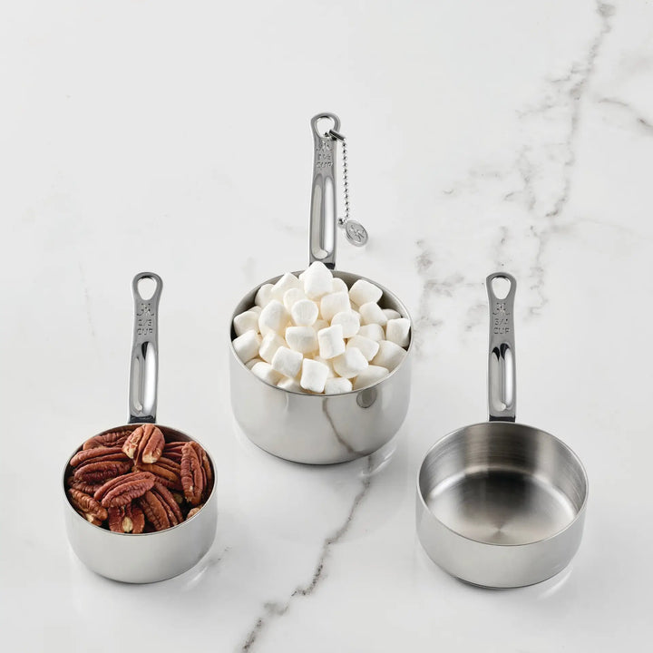 Three stainless steel measuring cups from the Hestan Provisions® Odd-Size Measuring Cups Set, 3-Piece are displayed on a white marble surface. The set includes a left cup filled with pecans, a middle cup holding mini marshmallows, and an empty right cup. Each Hestan Provisions® measuring cup features precise measurement markings on the handle for effortless accuracy.