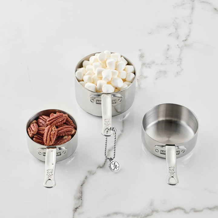 Three measuring cups from the Odd-Size Measuring Cups Set, 3-Piece by Hestan Provisions® are arranged on a marble surface. One cup from this premium stainless steel set is filled with pecans, another holds mini marshmallows, and the third remains empty. A small metal tag on a chain dangles from one of the handles, accentuating their elegant design.