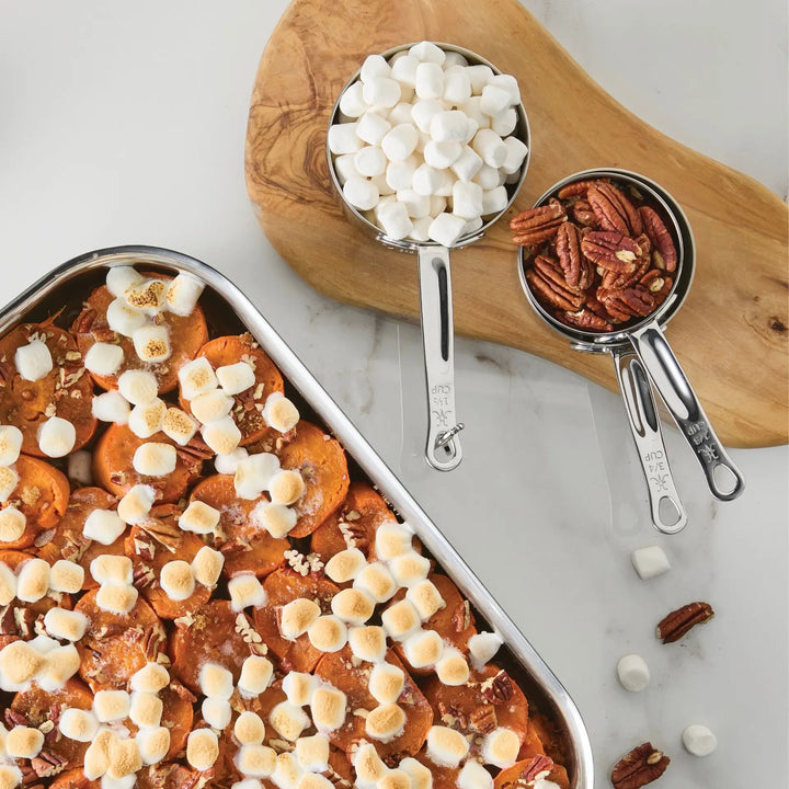 A baking dish of sweet potatoes topped with toasted mini marshmallows and pecans sits next to a wooden board holding the Hestan Provisions® Odd-Size Measuring Cups Set, 3-Piece, each cup filled with marshmallows and pecans on a white surface, embodying the precision of Provisions Chef’s Tools.