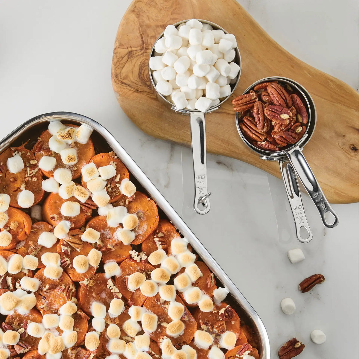 A baking dish of sweet potatoes topped with toasted mini marshmallows and pecans sits next to a wooden board holding the Hestan Provisions® Odd-Size Measuring Cups Set, 3-Piece, each cup filled with marshmallows and pecans on a white surface, embodying the precision of Provisions Chef’s Tools.
