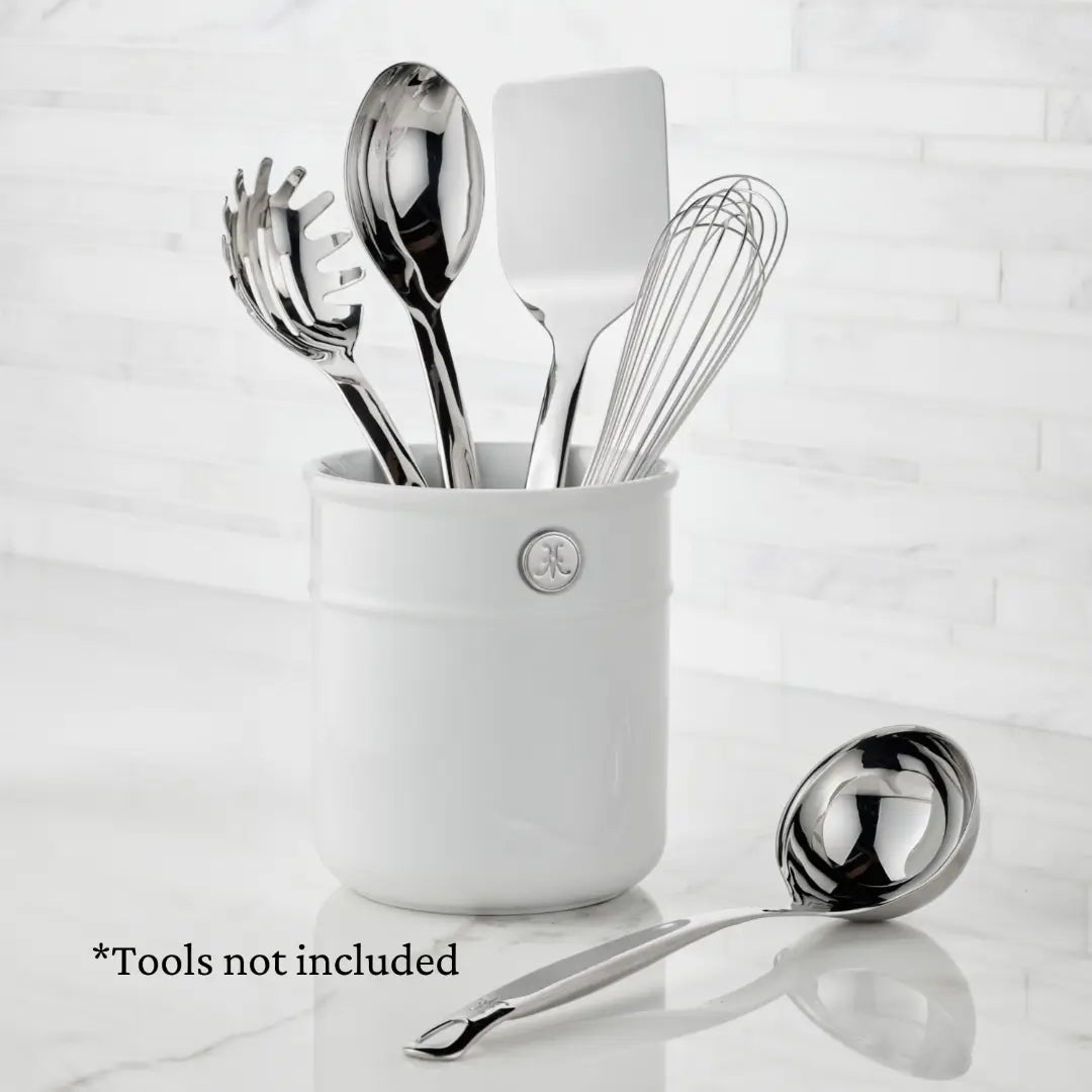 A white Porcelain Bain Marie by Hestan Provisions holds four essentials: pasta server, spatula, slotted spoon, and whisk. A ladle is beside it on a pristine surface. Text reads *Tools not included. Ideal for displaying your Hestan Culinary tools.