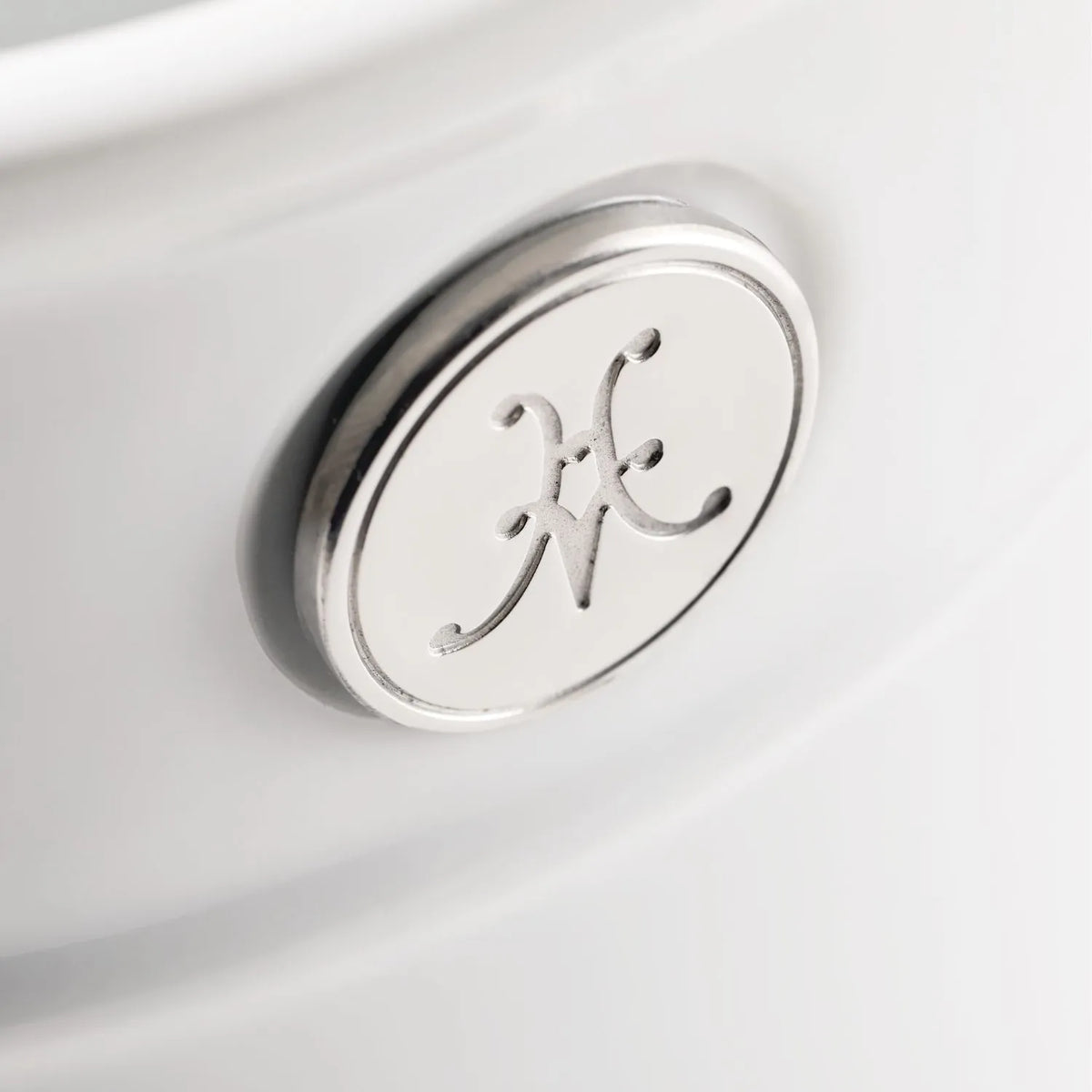 Close-up of a circular emblem with an intertwined letter design on a sleek, pale surface. Made from OvenBond Tri-ply Epicurean Stainless Steel, its polished, reflective finish enhances the sophisticated background. Brand: Hestan Provisions.