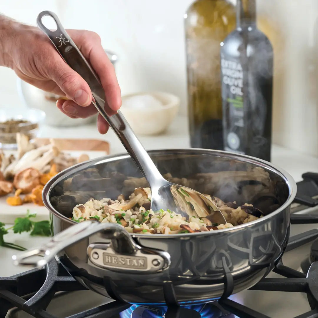 A hand stirs pasta and vegetables in a Hestan Culinary stainless steel pan on a gas stove, using the 13-inch Deep Solid Spoon by Hestan Provisions. Nearby are mushrooms, herbs, and olive oil as steam rises from this culinary masterpiece in progress.