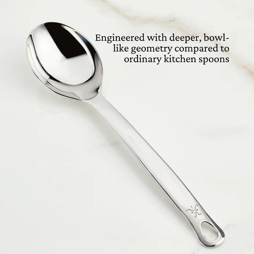 A Hestan Provisions Deep Solid Spoon, 13-inch with a stainless steel design and engraved handle rests on a white marble surface. Text highlights its unique geometry compared to regular kitchen spoons.