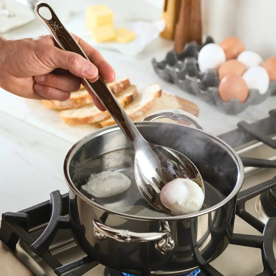 A hand uses a Hestan Provisions Deep Slotted Spoon, 13-inch, to lift a poached egg from a pot of boiling water on the stove. In the background, slices of bread, butter, and eggs in a carton rest on the countertop.