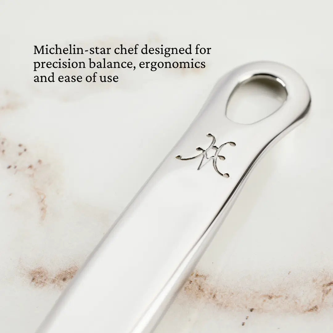 Close-up of a stainless steel utensil handle with an engraved symbol. On a light textured background, text reads: Michelin-star chef designed for precision balance, ergonomics, and ease of use—a hallmark of Chef’s Tools by Hestan Provisions. Product: Deep Slotted Spoon, 13-inch.”.