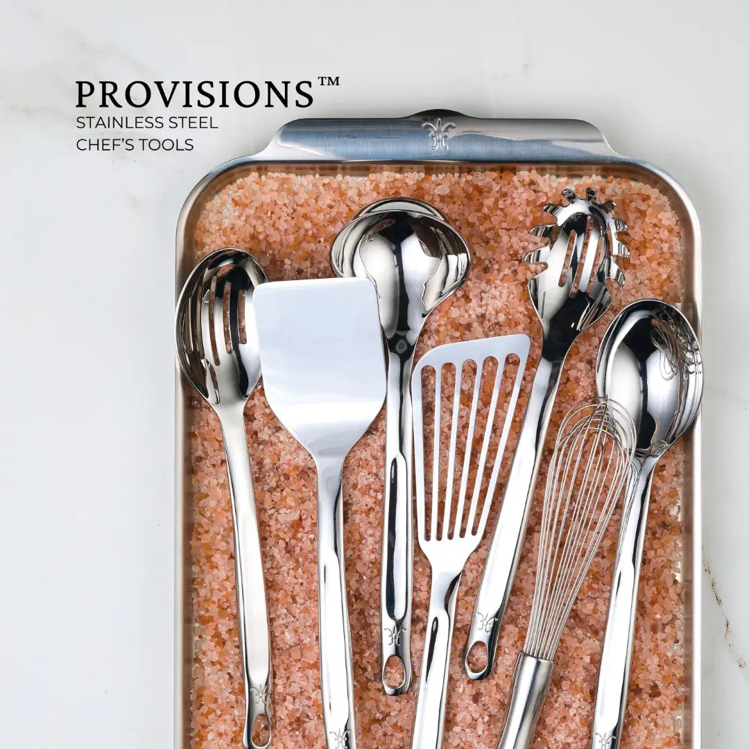 Hestan Provisions Chef’s Tools, featuring a 13-inch Deep Slotted Spoon, are elegantly arranged on a pink salt-filled tray. The brand and description are displayed in the upper left corner alongside the utensils, including spoons, spatulas, and a whisk.
