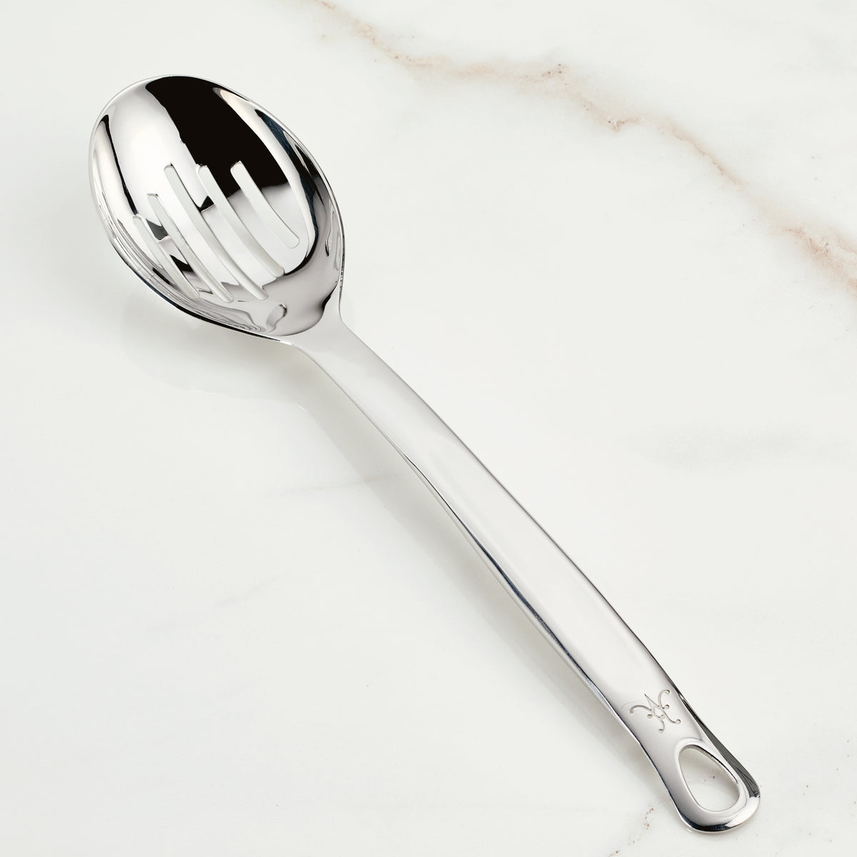 A shiny Hestan Provisions Deep Slotted Spoon, 13-inch, sits on a light marble surface. With decorative handle engravings and even slot spacing, it exemplifies expert craftsmanship and is essential for efficient liquid draining in any kitchen.