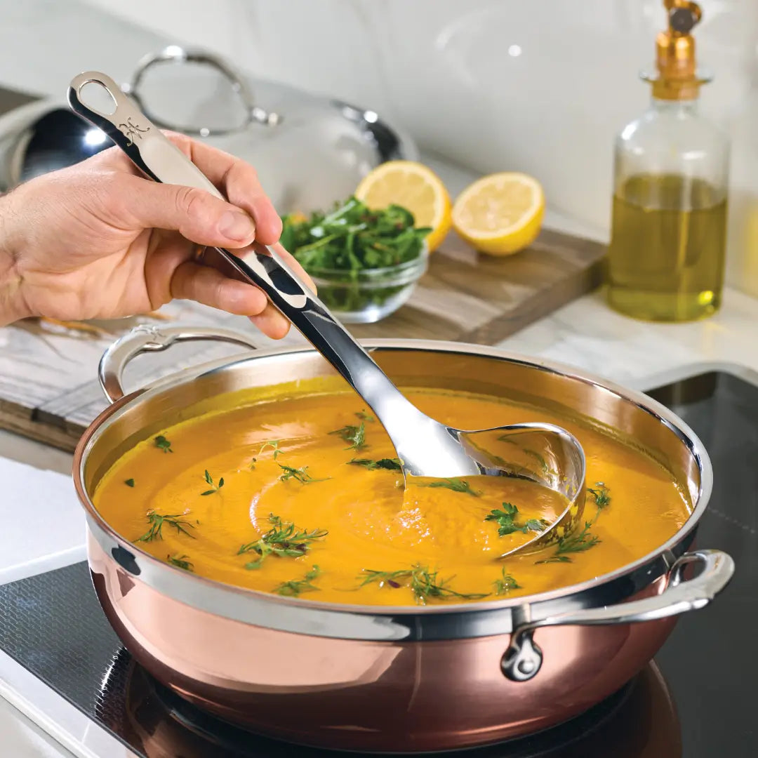 A hand stirs a pot of orange soup with Hestan Provisions 5-ounce ladle. Garnished with herbs, the soup is accompanied by a halved lemon, a small bowl of greens, and an olive oil bottle on a wooden board—arranged like Provisions Chef’s Tools from Hestan Culinary.