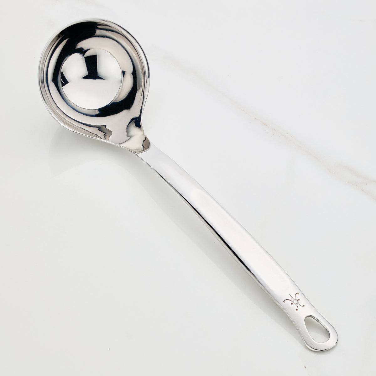 A Hestan Provisions Ladle, 5-ounce, with a shiny finish is elegantly placed on a white surface. It features a deep, rounded bowl and a long handle with a hole at the end for convenient hanging.
