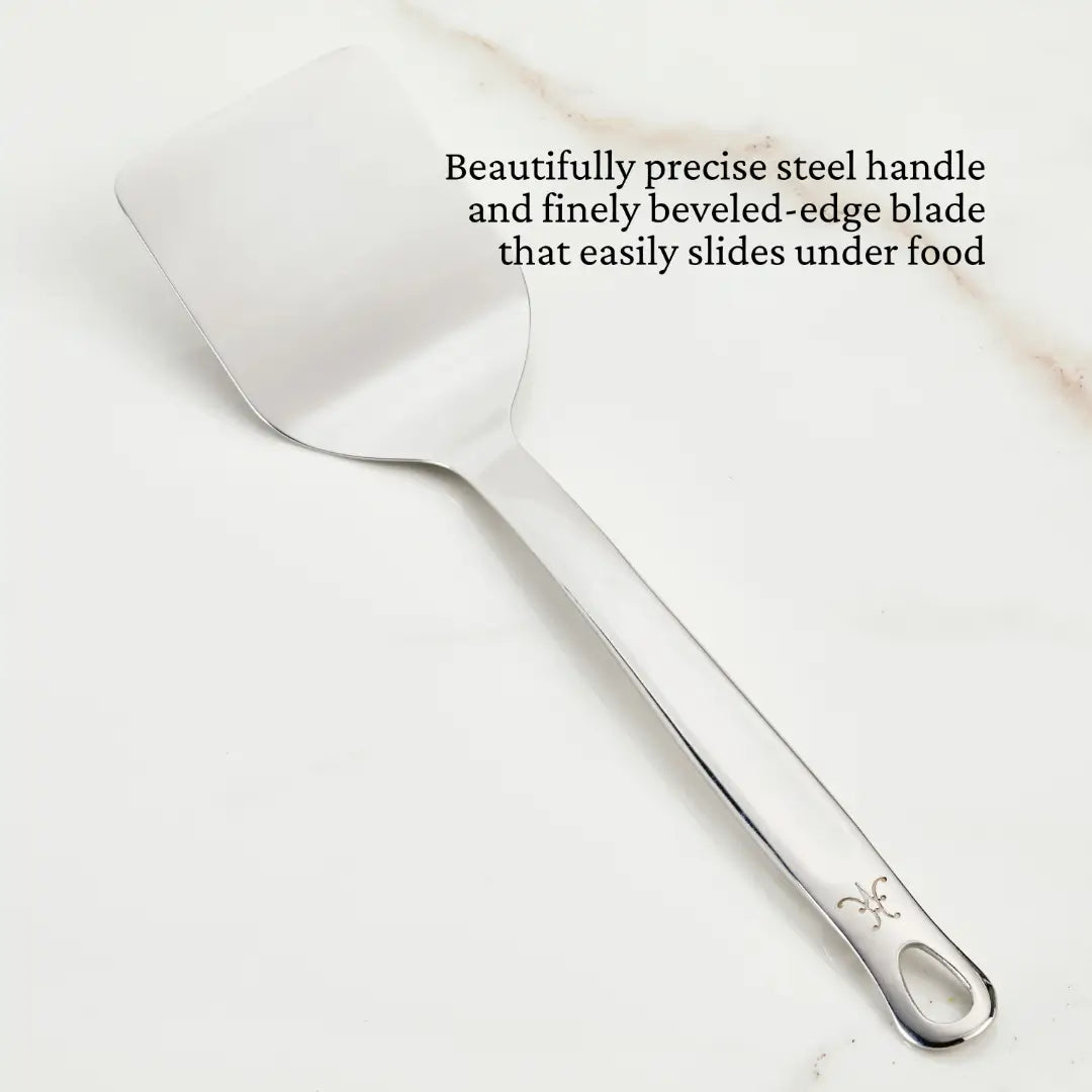 The Hestan Provisions 13-inch Solid Turner, ideal for chefs tools, boasts a flat, beveled-edge blade and a sleek handle. It sits gracefully on a white surface, emphasizing the outstanding design of its handle and blade.
