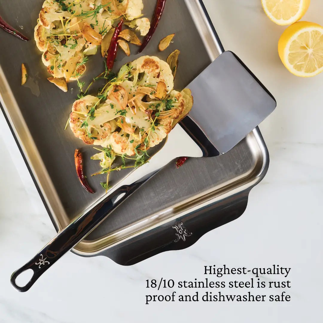 A stainless steel baking pan from Hestan Culinary holds roasted cauliflower with herbs and almonds, accompanied by a Solid Turner from Hestan Provisions. Nearby are lemon halves and red chili peppers. Rust-proof, dishwasher safe 18/10 stainless steel of the highest quality.