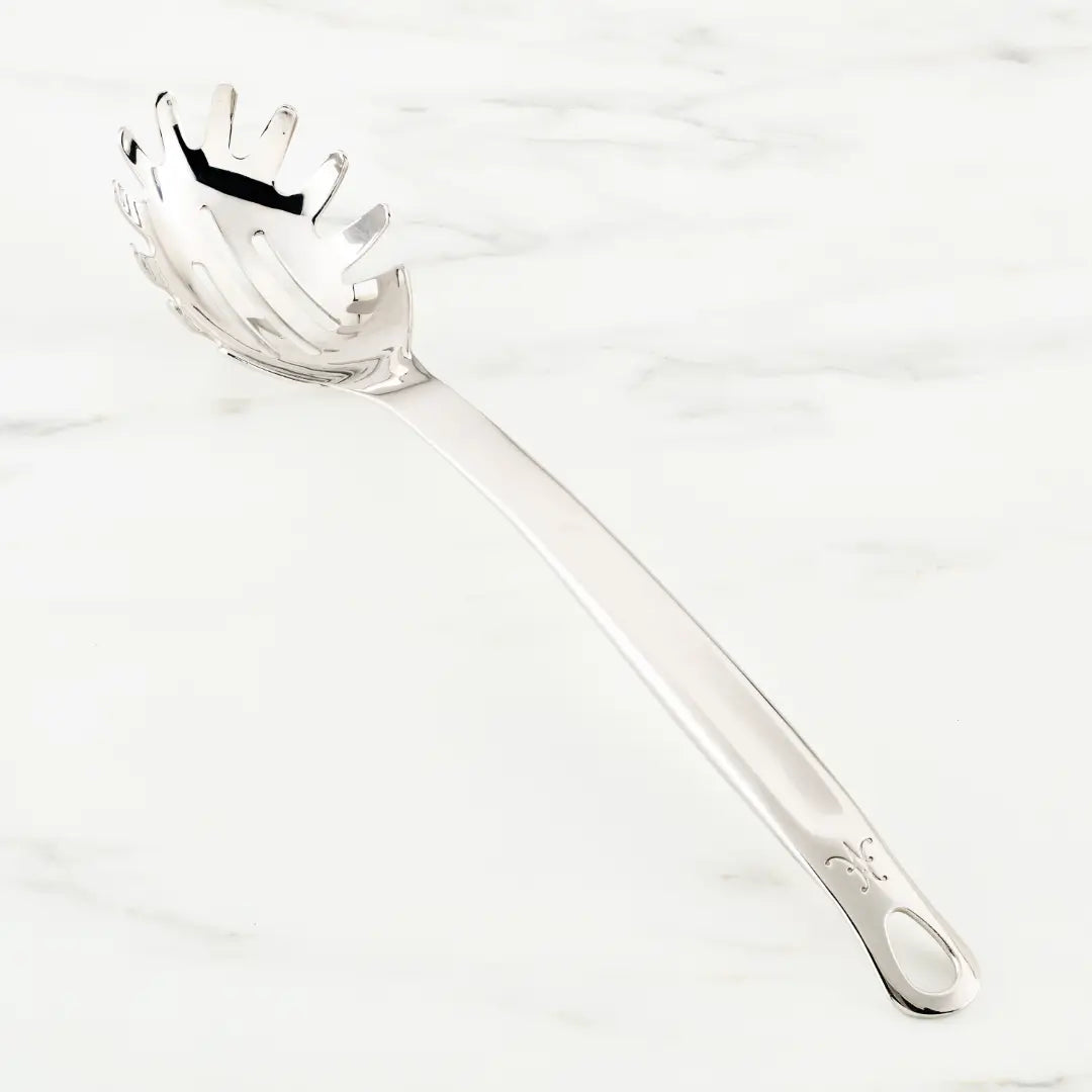 The Hestan Provisions Pasta Fork, 13-inch, made from 18/10 stainless steel, features a long handle with a decorative design and pronged head for easy pasta gripping. Displayed on light marble, it showcases the Provisions Chef’s Tools collection’s meticulous craftsmanship.