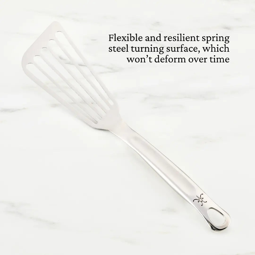 A Hestan Provisions Fish Turner, 13-inch, with a slotted, flexible stainless steel surface rests elegantly on a white marble countertop. The chef’s tool includes a hanging hole near the handles end and features simple decorative engraving.