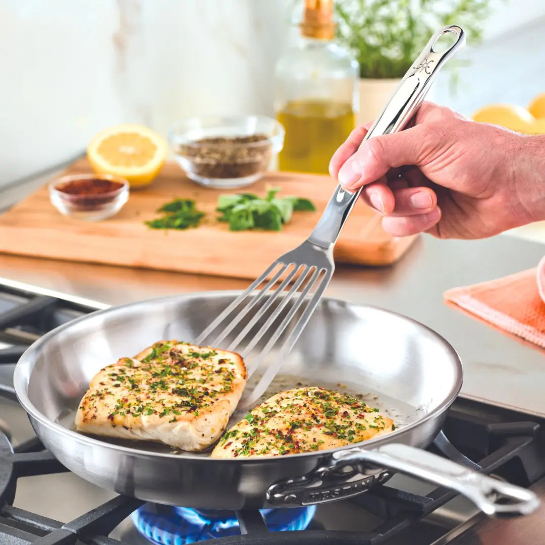A hand with a Hestan Provisions Fish Turner, 13-inch, expertly flips seasoned fish fillets in a stainless steel pan over a gas stove. In the background are herbs on a cutting board, lemon half, spices in a bowl, and olive oil. The flame flickers under the Hestan Culinary pan.