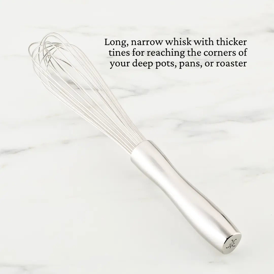 A Hestan Provisions French Whisk with thick, stainless steel tines and a sturdy handle lies on a light marble surface, praised for its ability to reach corners in deep cookware.