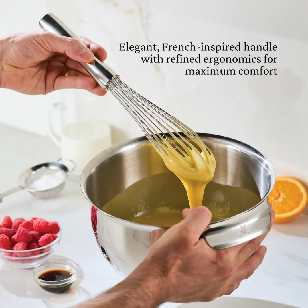 A person whisks yellow batter in a Hestan Provisions Chef’s Tools stainless steel bowl. Nearby lie a spoon with white powder, raspberries, half an orange, and a measuring cup with dark liquid. Text reads Elegant French Whisks by Hestan Provisions offer refined ergonomics for maximum comfort.