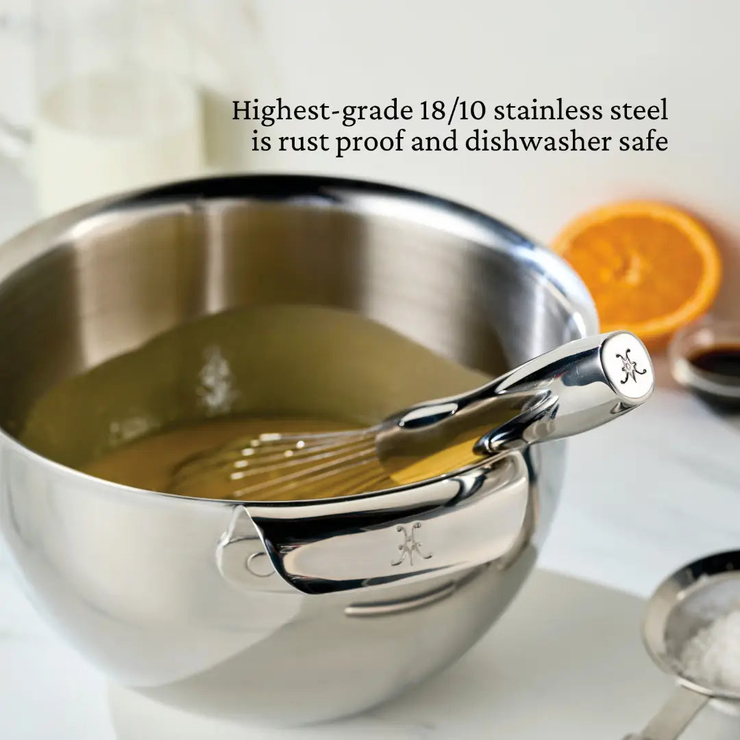 A Hestan Provisions French Whisk rests in a stainless steel bowl with a yellow liquid. In the background are an orange slice, chefs tools, and a glass jug. Text reads: 18/10 stainless steel is rust-proof and dishwasher safe.