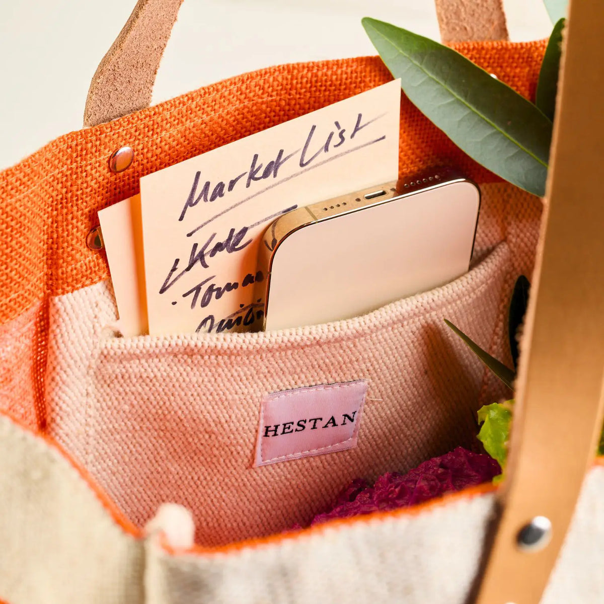 A close-up of the Hestan Market Tote in Citra Orange from Accessories reveals its durable luxury. The orange and beige bag holds a handwritten market list, a smartphone, and green leaves. Inside pocket features HESTAN label with kale and tomatoes elegantly penned on the list.