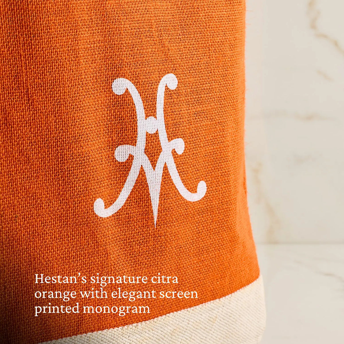 Close-up of the Hestan Market Tote in Citra Orange, showcasing a white monogram and bottom border, on light marble. Text: Hestans signature citra orange with elegant screen-printed monogram.