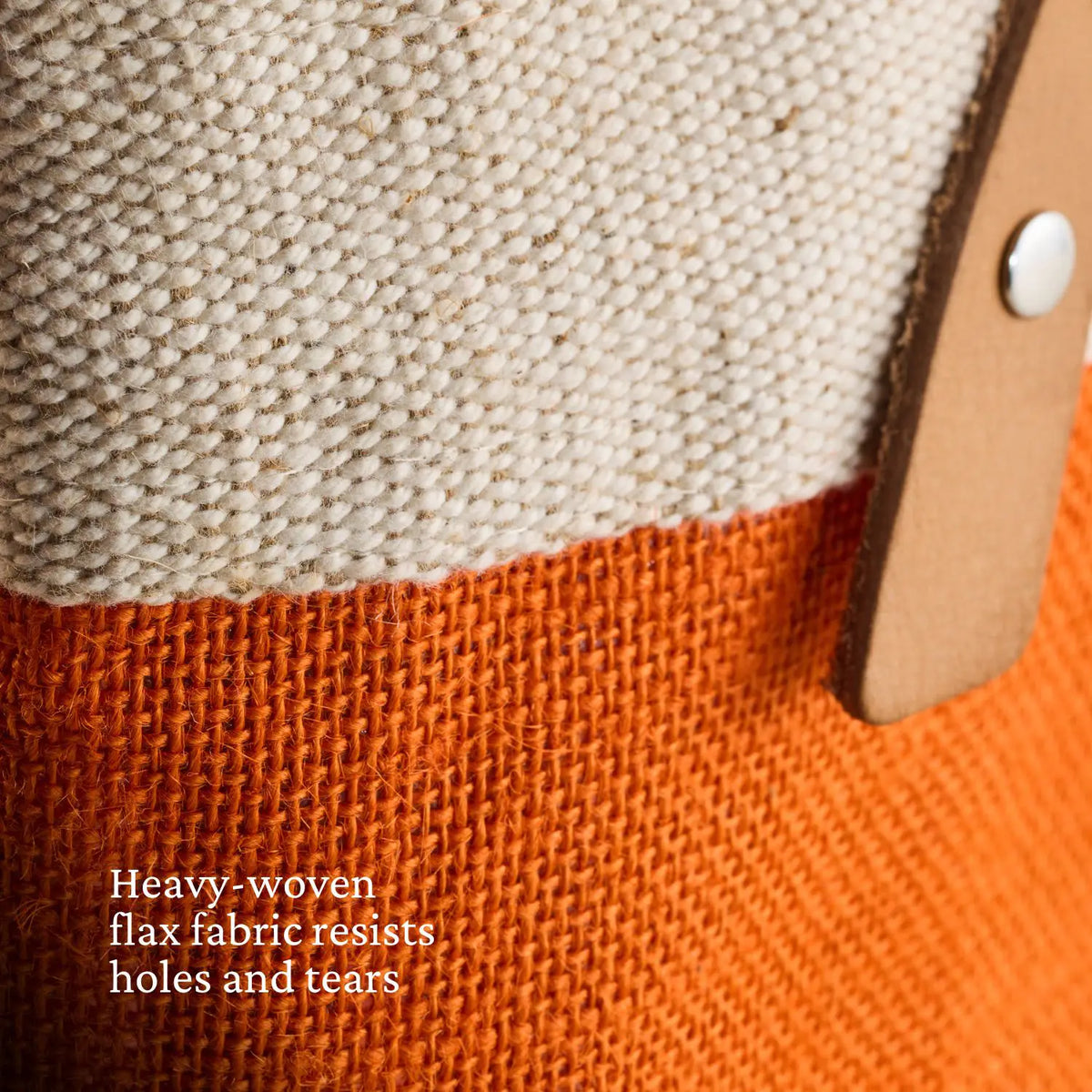 Close-up of heavy woven fabric: beige top, orange bottom. A tan leather strap with silver rivet on the right. Text reads, Accessories Hestan Market Tote, Citra Orange: Durable luxury flax resists holes and tears.