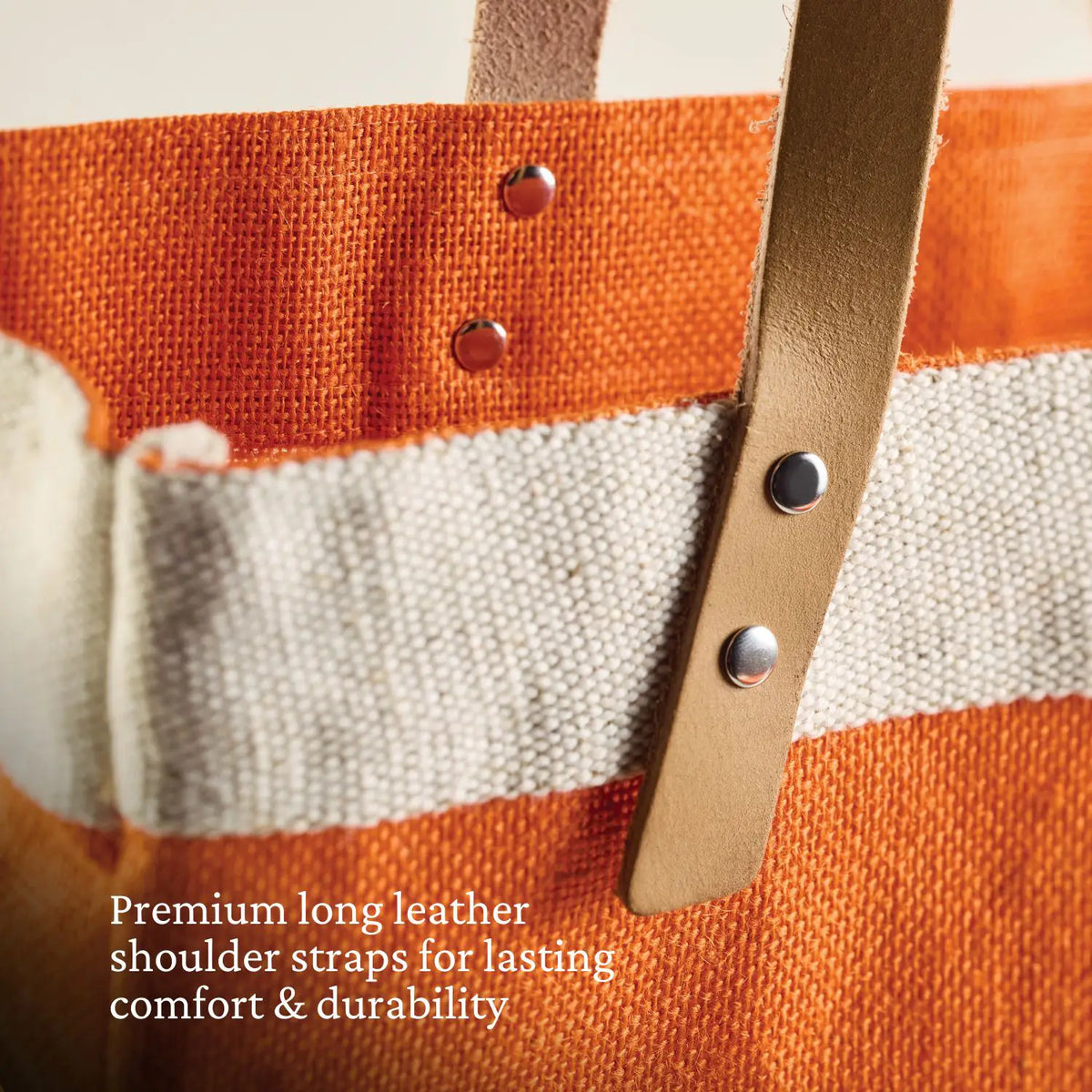 Close-up of the Hestan Market Tote, Citra Orange by Accessories: an orange fabric bag with a beige stripe and leather shoulder straps adorned with metal rivets. Text: Premium long leather shoulder straps for durable luxury & lasting comfort.