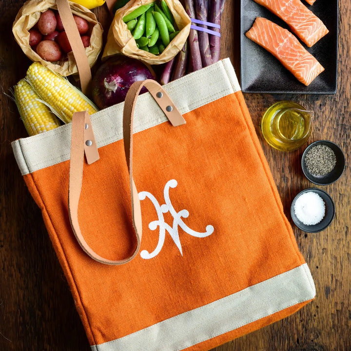 A Hestan Market Tote in Citra Orange by Accessories rests on a wooden surface, featuring brown handles and a white monogram, surrounded by corn, potatoes, purple beans, peas, red onion, raw salmon, olive oil, black pepper, and salt.
