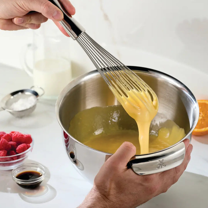 Hands whisk thick yellow batter in a Hestan Provisions Stainless Steel Mixing Bowl. Nearby, kitchenware includes a small bowl of raspberries, a cup of liquid, cocoa powder or coffee, half an orange, and a cup of sugar on the countertop.